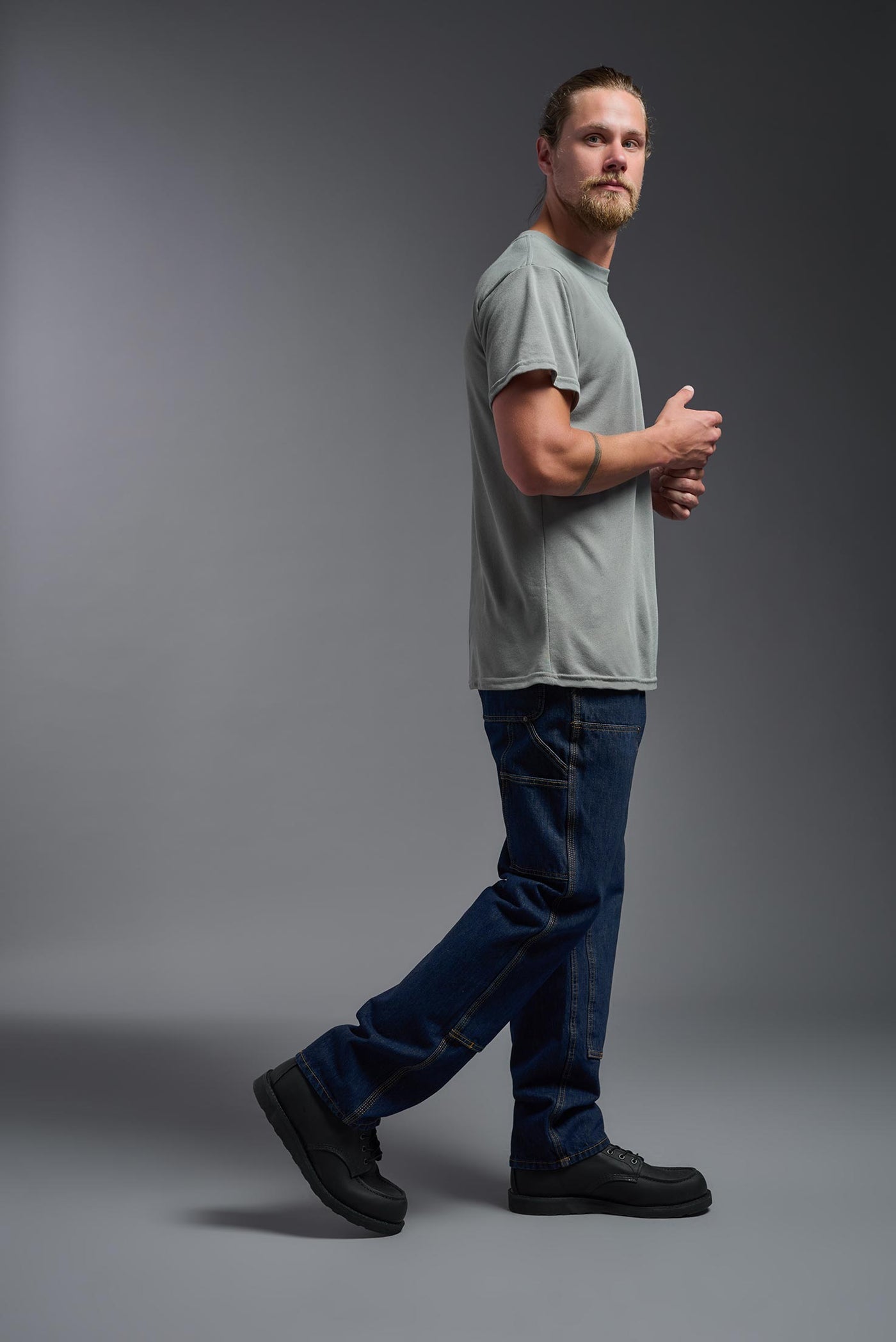 A male caucasian model with a beard wearing the men's DuroSoft Outdoor Tee in the color army gray on a gray background, made out of 100% filament polyester that feels like soft cotton, showing the modern fit from a side view. This shirt is anti-snagging, quick-drying and moisture-wicking and is made in the USA. #color_army-gray