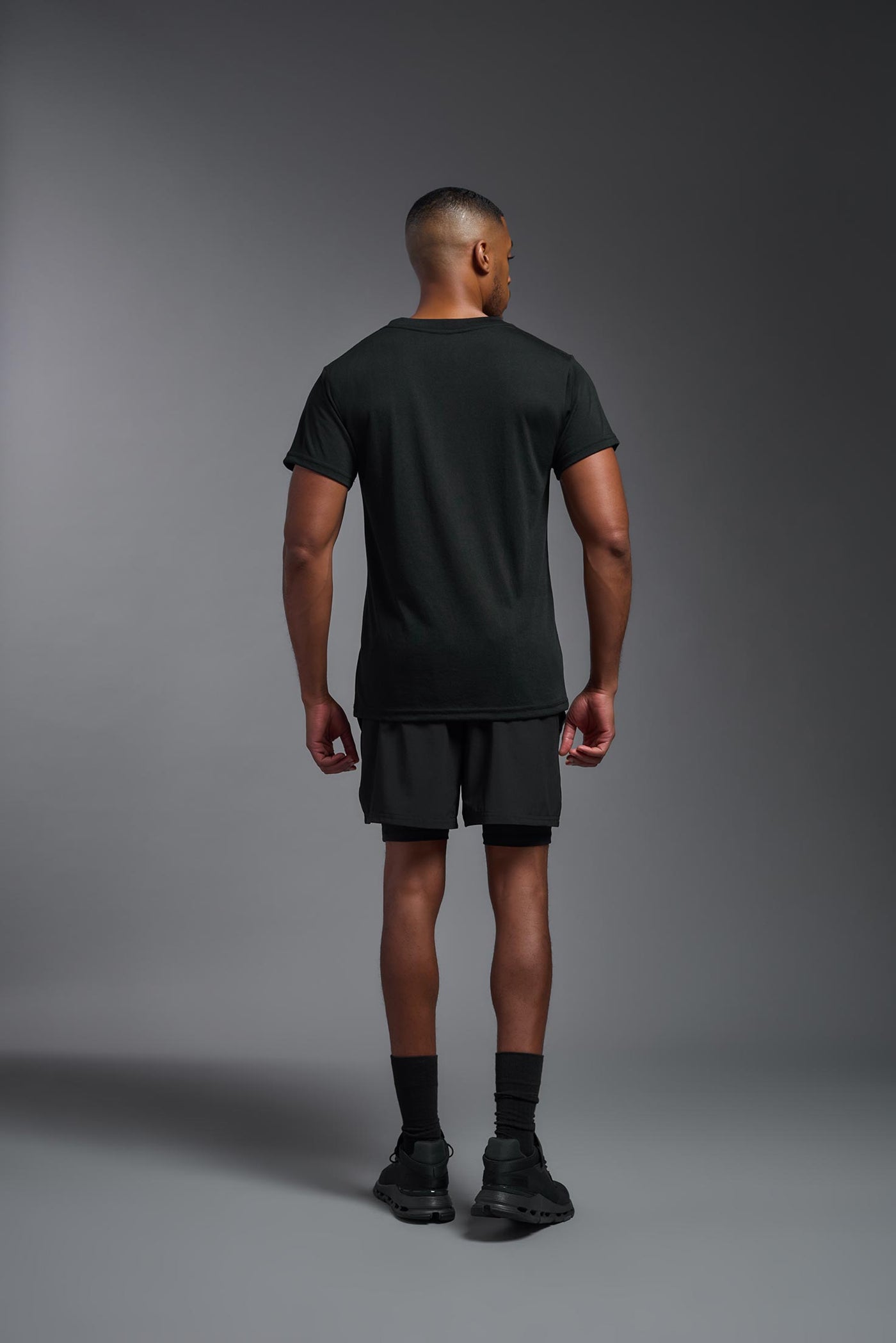 A male African American model with a beard wearing the men's DuroSoft Outdoor Tee in the color black on a gray background, made out of 100% filament polyester that feels like soft cotton, showing the modern fit from a back view. This shirt is anti-snagging, quick-drying and moisture-wicking and is made in the USA. #color_black