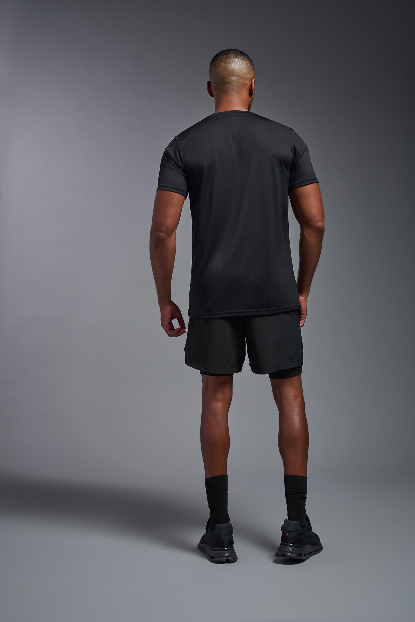 A male African American model with a beard wearing the men's EcoTek™ Recycled Performance Tee in the color black, made out of 100% recycled polyester REPREVE®, showing the modern fit from a back view. This shirt features a mechanical stretch, quick-dries, wicks moisture and is made in the USA. #color_black