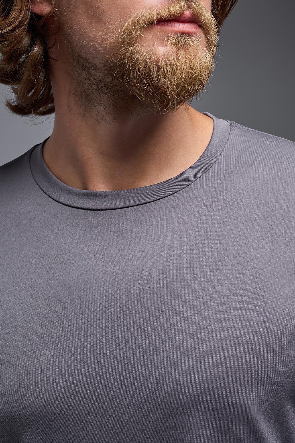 A male blonde model with a beard wearing the men's EcoTek™ Recycled Performance Tee in the color charcoal, made out of 100% recycled polyester REPREVE®, showing the modern fit from a collar close-up view. This shirt features a mechanical stretch, quick-dries, wicks moisture and is made in the USA. #color_charcoal