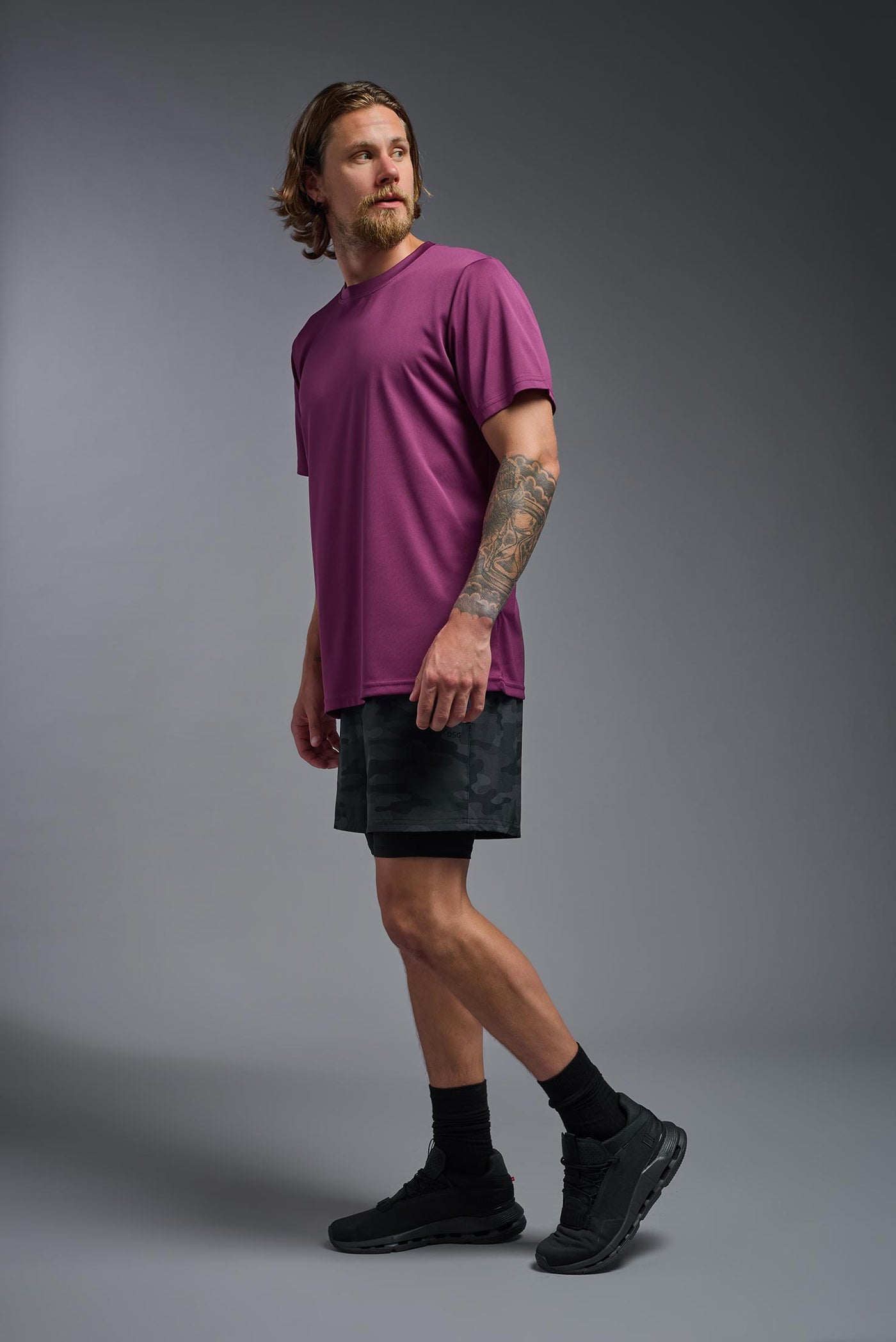 A blonde, male model with a beard wearing the men's EcoTek™ Recycled Performance Tee in the color merlot, made out of 100% recycled polyester REPREVE®, showing the modern fit from a side view. This shirt features a mechanical stretch, quick-dries, wicks moisture and is made in the USA. #color_merlot