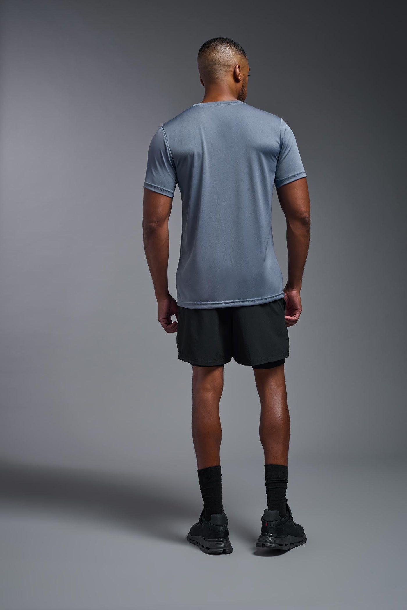 An African American, male model with a beard wearing the men's EcoTek™ Recycled Performance Tee in the color tahoe blue, made out of 100% recycled polyester REPREVE®, showing the modern fit from a back view. This shirt features a mechanical stretch, quick-dries, wicks moisture and is made in the USA. #color_tahoe