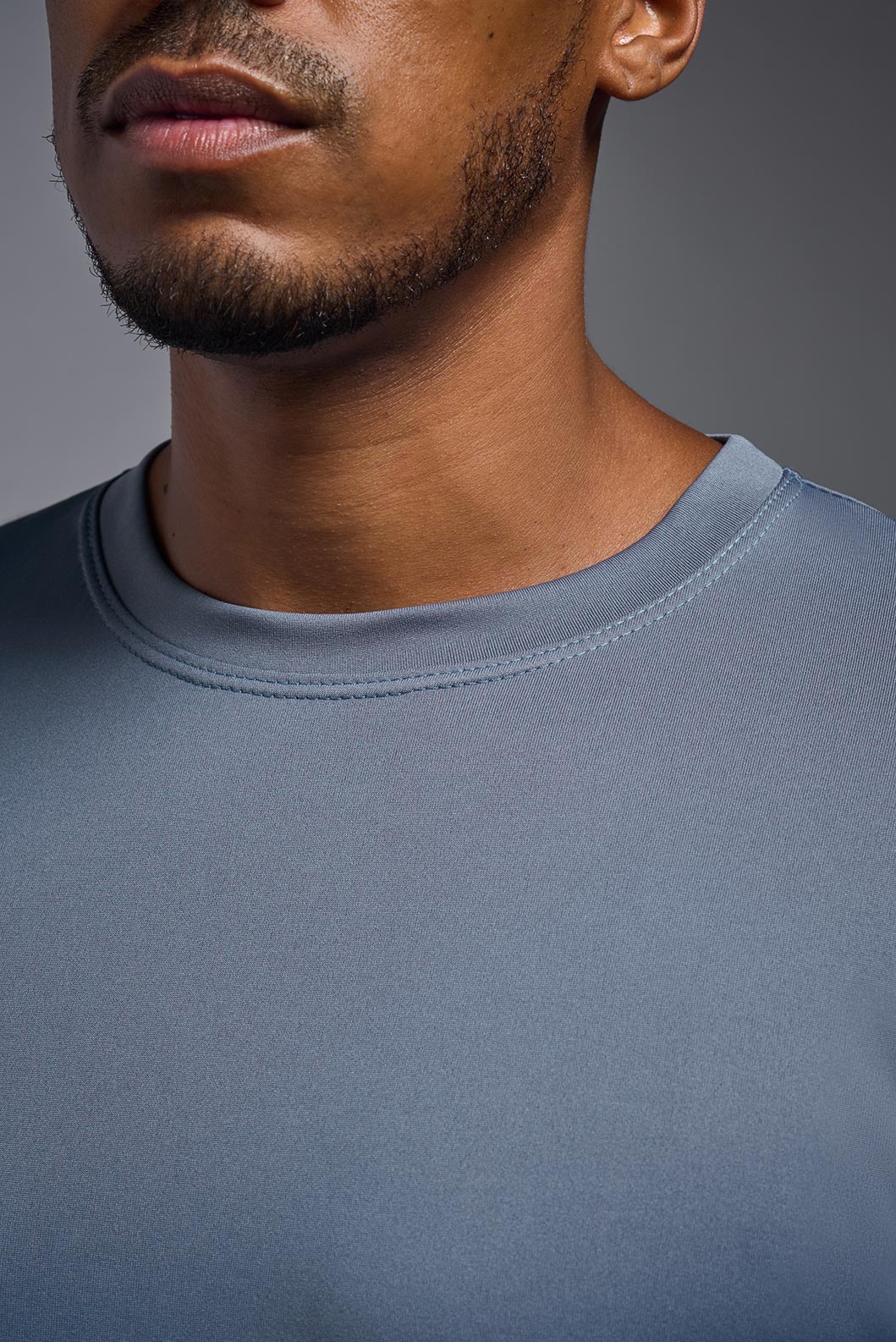 An African American, male model with a beard wearing the men's EcoTek™ Recycled Performance Tee in the color tahoe blue, made out of 100% recycled polyester REPREVE®, showing the modern fit from a close-up collar view. This shirt features a mechanical stretch, quick-dries, wicks moisture and is made in the USA. #color_tahoe