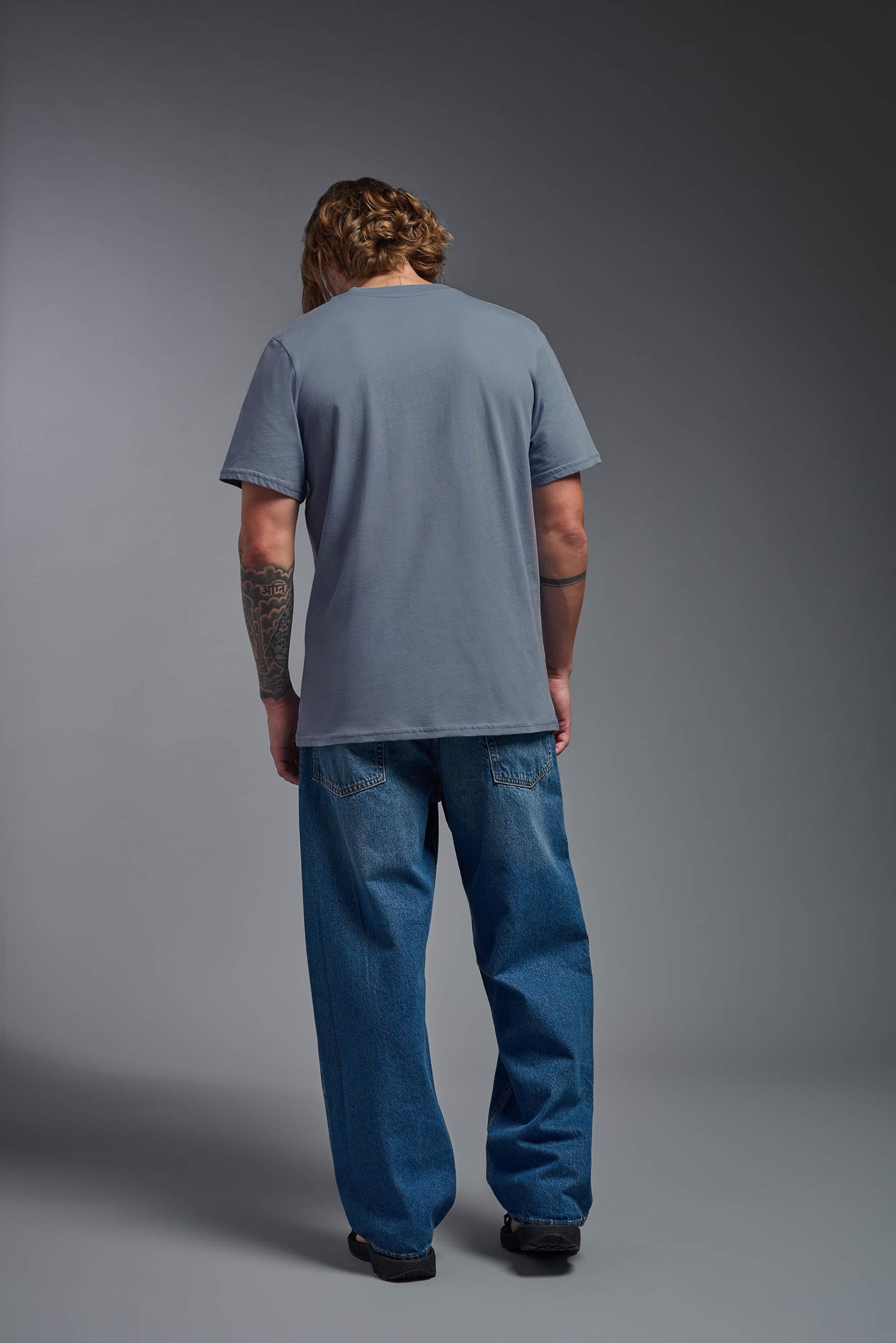A male model wearing the unisex Organic Soft Cotton T-shirt in the color canyon blue, made from 100% certified organic cotton, shot from a full body back view. This shirt is unisex and made in the USA. #color_canyon-blue
